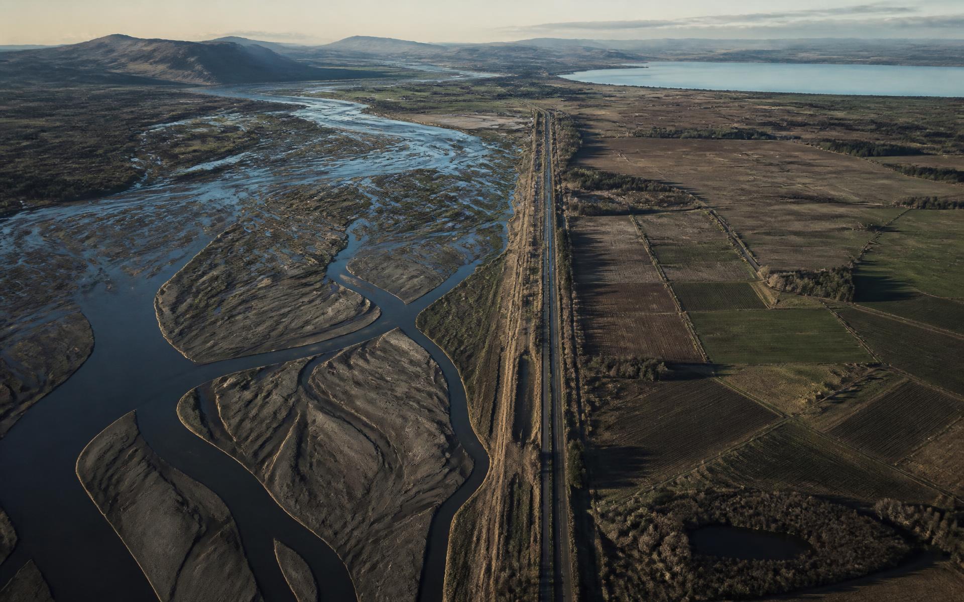 Aerial view of a river delta plain crossed by a highway and rail corridor toward distant terrain